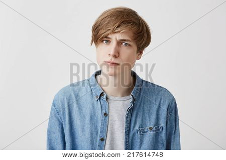 Portrait of young irritated caucasian male with fair hair and blue eyes looks with anger at camera, dissatisfied hear some comments, tries to control emotions and not speak out his mind in presence of many people and guests