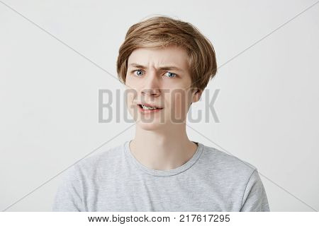 Grumpy irritated fair-haired young male being emotional and angry after offended by cruel man, recieves bad words, isolated against gray background. Bewilderment concept