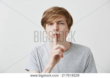 Portrait of angry irritated fair-haired male asks keep confidential information in secret, annoyed with gossips, demands quiet, says: Hush, stop talking. Caucasian guy makes silence gesture