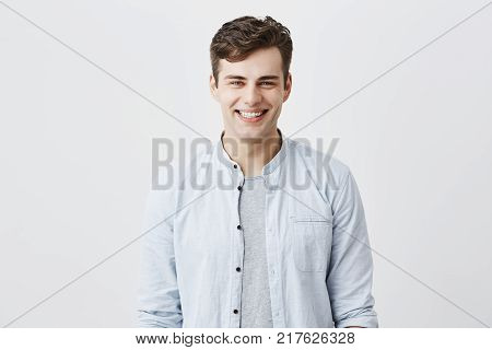 Handsome attractive young man dressed in light blue shirt over t-shirt with dark hair and appealing blue eyes looknig at camera, smiling broadly, demonstrating white teeth, being happy and pleased, posing indoors. Positive emotions