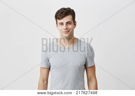 Stylish young caucasian man with appealing blue eyes smiling into camera having pleased expression being glad to meet his girlfriend. Hipster guy dressed in gray t-shirt grinning having cheerful look. Positive emotions