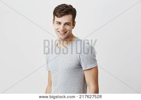 Positive european guy with blue eyes dressed casually having pleasant smile showing his perfect white teeth having good mood after party and meeting with his friends. Cheerful good-looking male model posing against gray background.
