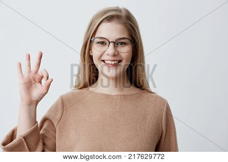 Young European blonde female showing OK-gesture with her fingers. Happy girl in brown sweater and eyeglasses broadly smiling. Her happy face proves everything goes according to plan
