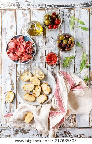 Ingredients for making tapas or bruschetta. Crusty bread, ham prosciutto, sun dried tomatoes, olive oil, olives, pepper, greens on plates over white plank wooden table. Top view with space