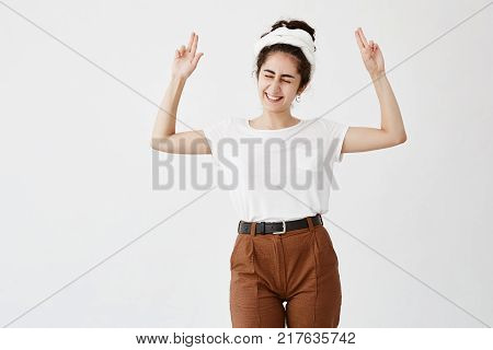 Excited overjoyed woman with dark and wavy hair in bun points at copy space, closing her eyes, smiles broadly, advertisizes. Happy young female in white t-shirt poses against white wall with copy area for your promotional text