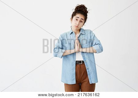Sorrorful religious dark-haired young woman holding hands in prayer hoping for fortune. Religion, spirituality concept. Sad student girl in denim shirt begging for mark