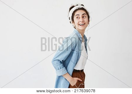 Pleasant looking lauging joyful female with wavy hair in bun, keeping hand in pocket, using mobile phone for communication with friends or lover, listening to pleasant music, having good mood.