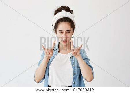 Positive gloomy young girl with dark and wavy hair in bun, wearing do-rag having fun, pouting lips, showing rock sign with both hands. Body language and gestures concept.