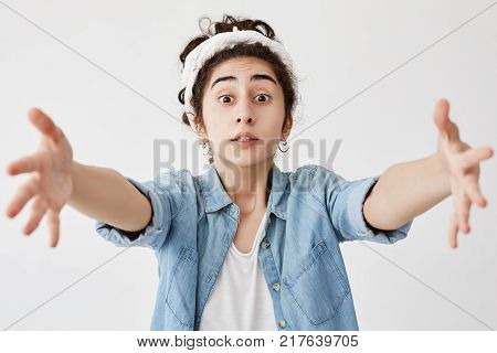 Studio shot of pretty girl with wide opened eyes wearing do-rag and denim shirt asking for something, stretching her arms to camera to get what she wants. Cute dark-haired young poses against white blank wall