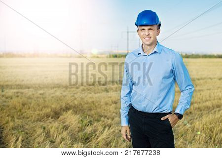 engineer or worker smiles in protective helmet holding a phone. Wheat fiels, power line, sunny day, blue sky.