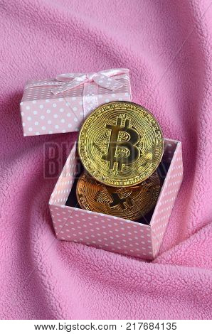 The golden bitcoin lies in a small pink gift box with a small bow on a blanket made of soft and fluffy light pink fleece fabric with a large number of relief folds