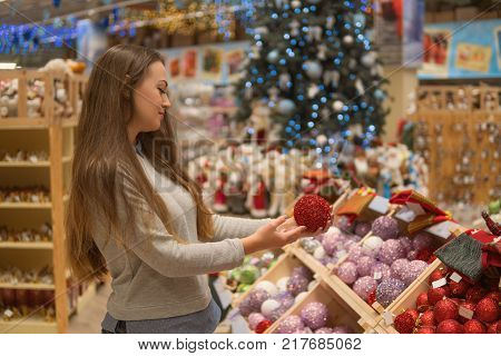 Cheerful happy customer girl choosing gifts for Christmas and New Year. She is taking red Christmas tree decoration balls. difficult decision. Various choice