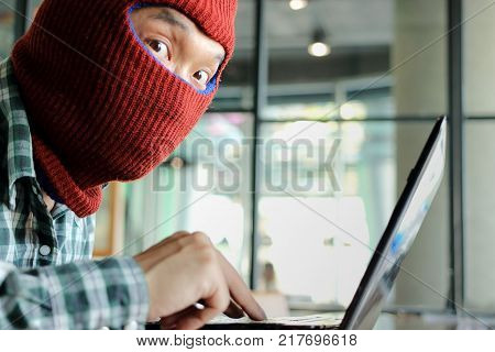 Masked hacker wearing a balaclava using laptop stealing important information data. Internet security and privacy crime concept. Selective focus and shallow depth of field.