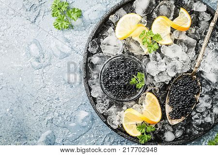 Black caviar in a glass bowl on ice with lemon close up over gray background, top view