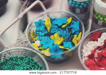 Ice cream treats and toppings sale. Rainbow sprinkles, flowers, non pareil and glitter cupcake decorations, bright yellow and blue. Closeup in plastic plates with spoons. Confectionery store