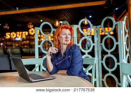 A woman, aged, red-haired, in a blue dress and glasses, looks away thoughtfully with glasses in her hand behind a stellite in a cafe