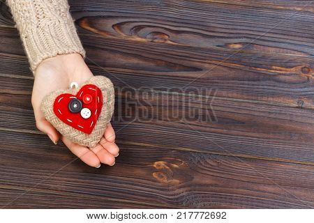 Woman's hands with beautiful accurate knitted red heart. White cozy knitted sweater on wooden background. Concept of giving help. Love concept with copyspace.