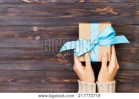 Woman hands give wrapped valentine or other holiday handmade present in paper with red blue and green ribbon. Present box decoration of gift on white wooden table top view with copy space.