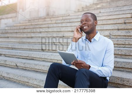 Happy black businessman working on tablet. African-american insuranse agent checking email on digital device, talking on mobile, sitting on the stairs outdoors in urban area
