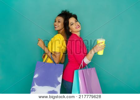 Happy multiethnic girlfriends with shopping bags and take away drink. Two happy excited smiling shopaholics at turquoise sudio background with copy space