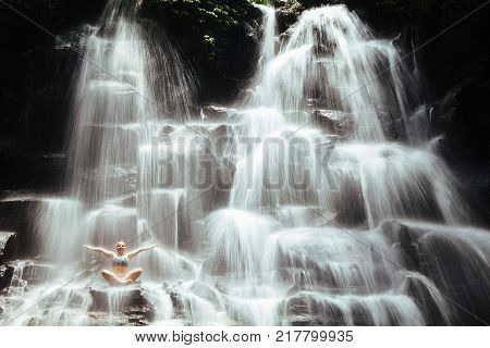 Travel in Bali jungle. Beautiful young woman sitting in yoga pose on rock under falling spring water enjoy tropic cascade waterfall. Nature day trip walking adventure fun on family summer vacation