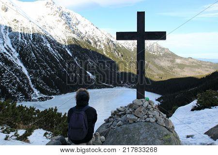 A young man sits near a large cross on a background of mountains