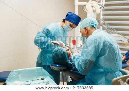 Male Old Professional Dentist Surgeon In Uniform And Female Assistant Helps To Performing Operation