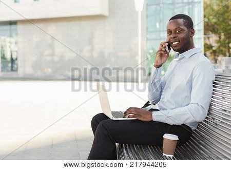 Happy black businessman working on laptop. African-american insuranse agent checking email on computer, talking on mobile, sitting on the bench outdoors near modern office center