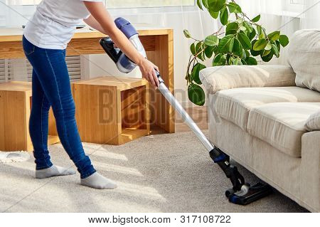Portrait Of Young Woman In White Shirt And Jeans Cleaning Carpet Under Sofa With Vacuum Cleaner In L