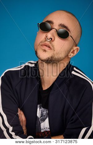Studio Shot Of A Young Tattoed Bald Man Posing Against A Blue Background. 90s Style.