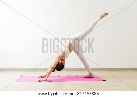 Young Female Practicing One Legged Downward Facing Dog Pose On Mat In Fitness Studio