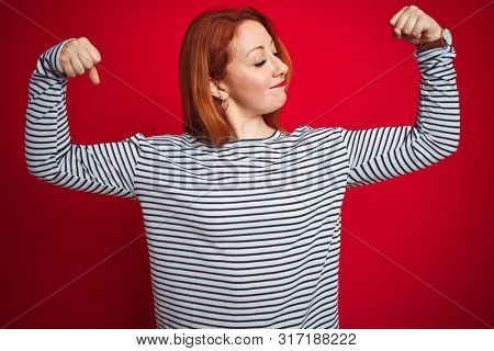 Young redhead woman wearing strapes navy shirt standing over red isolated background showing arms muscles smiling proud. Fitness concept.