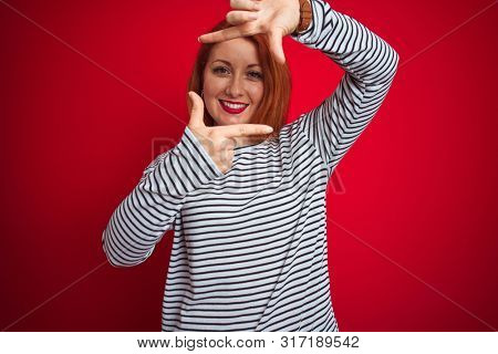 Young redhead woman wearing strapes navy shirt standing over red isolated background smiling making frame with hands and fingers with happy face. Creativity and photography concept.