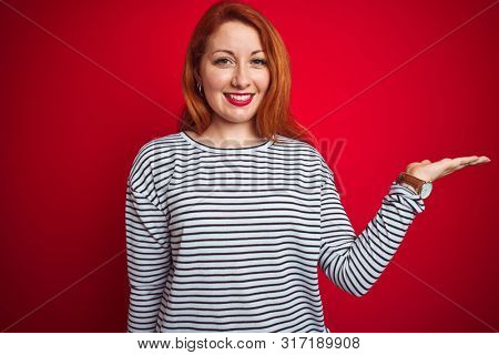 Young redhead woman wearing strapes navy shirt standing over red isolated background smiling cheerful presenting and pointing with palm of hand looking at the camera.