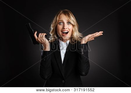 Portrait of a beautiful happy blonde haired business woman dressed in formal clothes standing isolated over black background, shooting with a gun