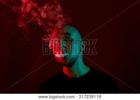 Close-up Studio Shot Of A Young Bald Guy Vaping, Blowing Out A Cloud Of Smoke On Red Background.