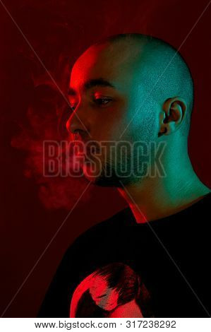 Close-up Studio Shot Of A Young Bald Guy Vaping, Blowing Out A Cloud Of Smoke On Red Background.