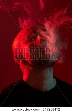 Close-up Studio Shot Of A Young Bald Guy Vaping, Blowing Out A Cloud Of Smoke On Red Background.