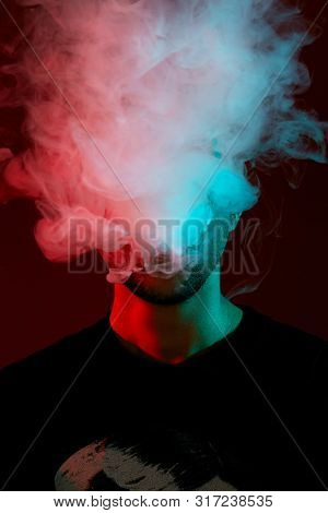Close-up Studio Shot Of A Young Bald Guy Vaping, Blowing Out A Cloud Of Smoke On Red Background.