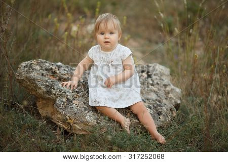 Cute Little Baby Girl Sitting On A Rock In Nature. Looking Into The Camera. The Concept Is Tendernes