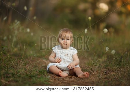 A Cute Little Baby Girl In A Light Dress Is Sitting On A Path In The Forest. Looking Into The Camera
