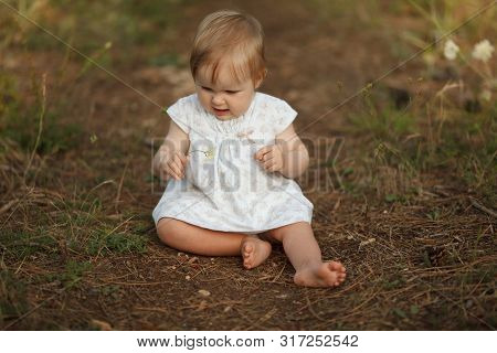 Cute Little Baby Girl In A Light Dress Sitting In Nature. Looks At Flower. The Concept Is Tenderness