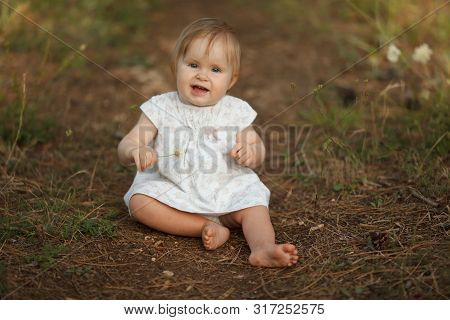 Beautiful Little Baby Girl In A Light Dress With A Flower In Nature. Looking At The Camera. The Conc