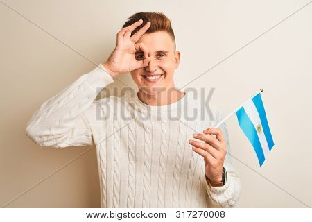 Young handsome man holding Argentinian Argentina flag over isolated white background with happy face smiling doing ok sign with hand on eye looking through fingers