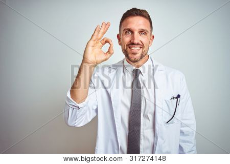 Young handsome doctor man wearing white profressional coat over isolated background smiling positive doing ok sign with hand and fingers. Successful expression.