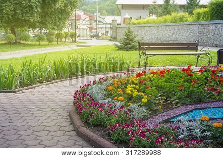 Round Flower Bed With Colorful Decorative Flowers And Plants, Garden Bench Stands On The Paving Tile