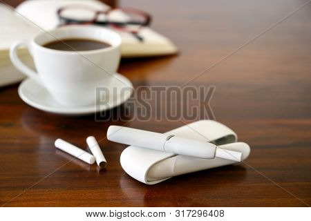 Heating Tobacco System, Coffee Cup And A Book On A Brown Table, E-cigarette With Tobacco Sticks That