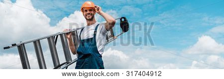 Panoramic Shot Of Cheerful Repairman Holding Ladder And Smiling Against Blue Sky With Clouds