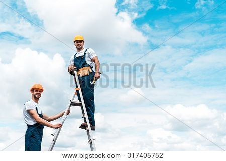 Happy Repairmen Standing On Ladder And Smiling Against Blue Sky With Clouds