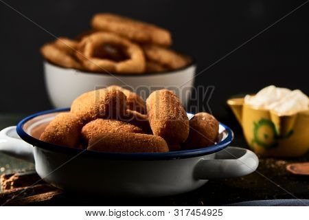 some spanish croquettes in a white and blue enamel plate on a table and some calamares a la romana, fried battered squid rings typical of spain, in the background, next to a mortar with aioli sauce